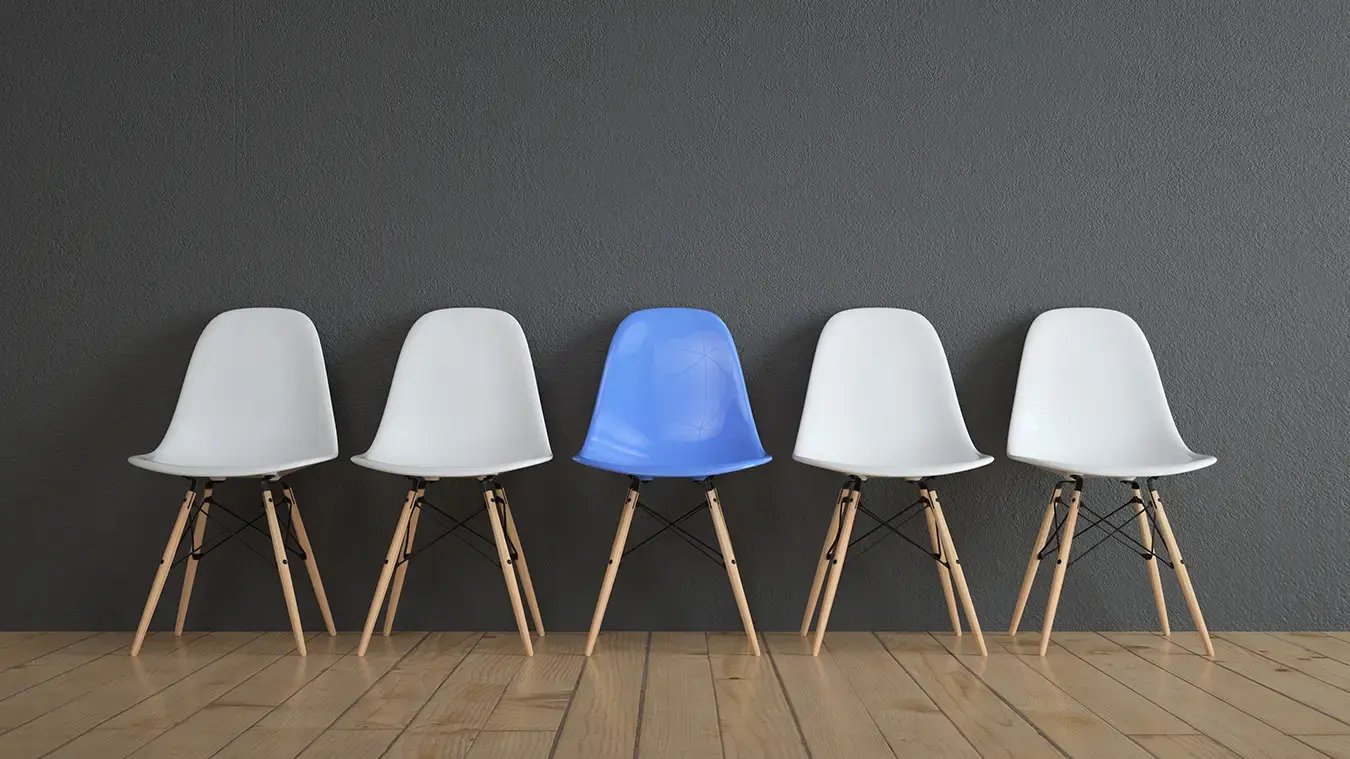 Five chairs against a black wall. One blue chair stands out among four white chairs.