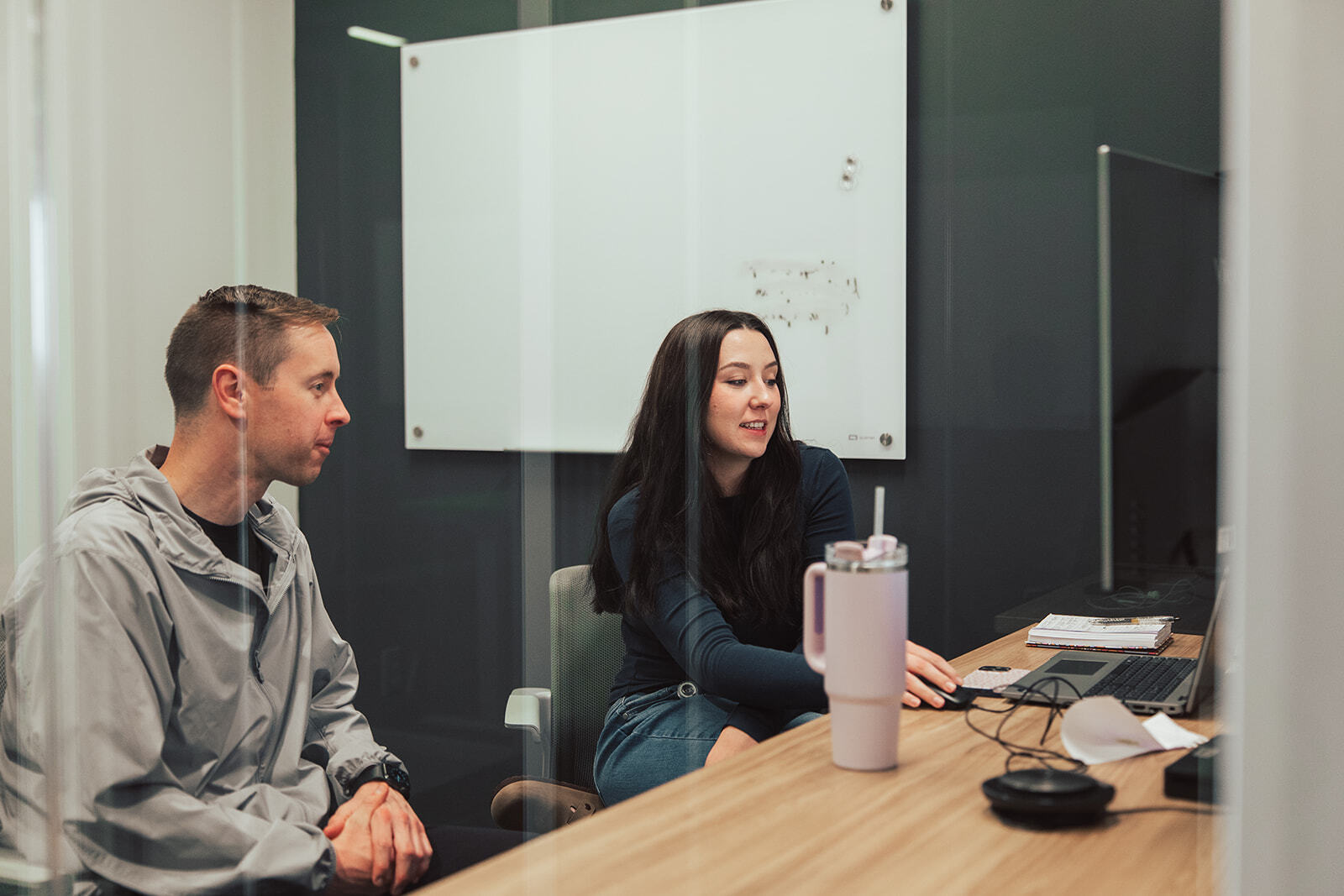 Two people sitting at a computer desk in an enclosed office having a conversation.